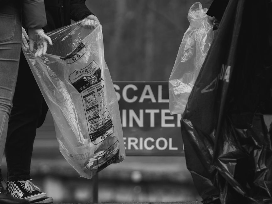 Two individuals are shown handling clear and black plastic rubbish bags. The person on the left, wearing jeans and checkerboard pattern shoes, is holding a transparent bag filled with mixed waste, inc