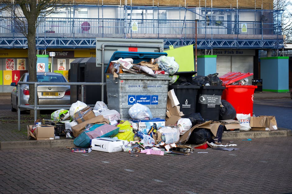 In the foreground, a large collection of overflowing rubbish and waste materials is gathered on a paved sidewalk, including black, green, and red plastic refuse bins, some of which are open with conte